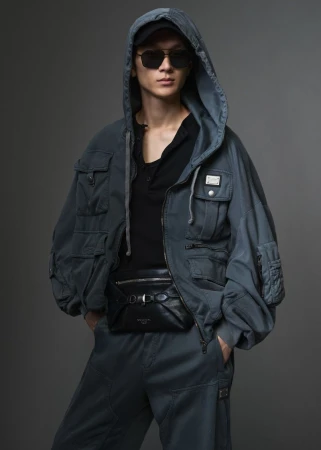 Model wearing a grey hooded utility jacket and sunglasses against a solid dark grey backdrop
