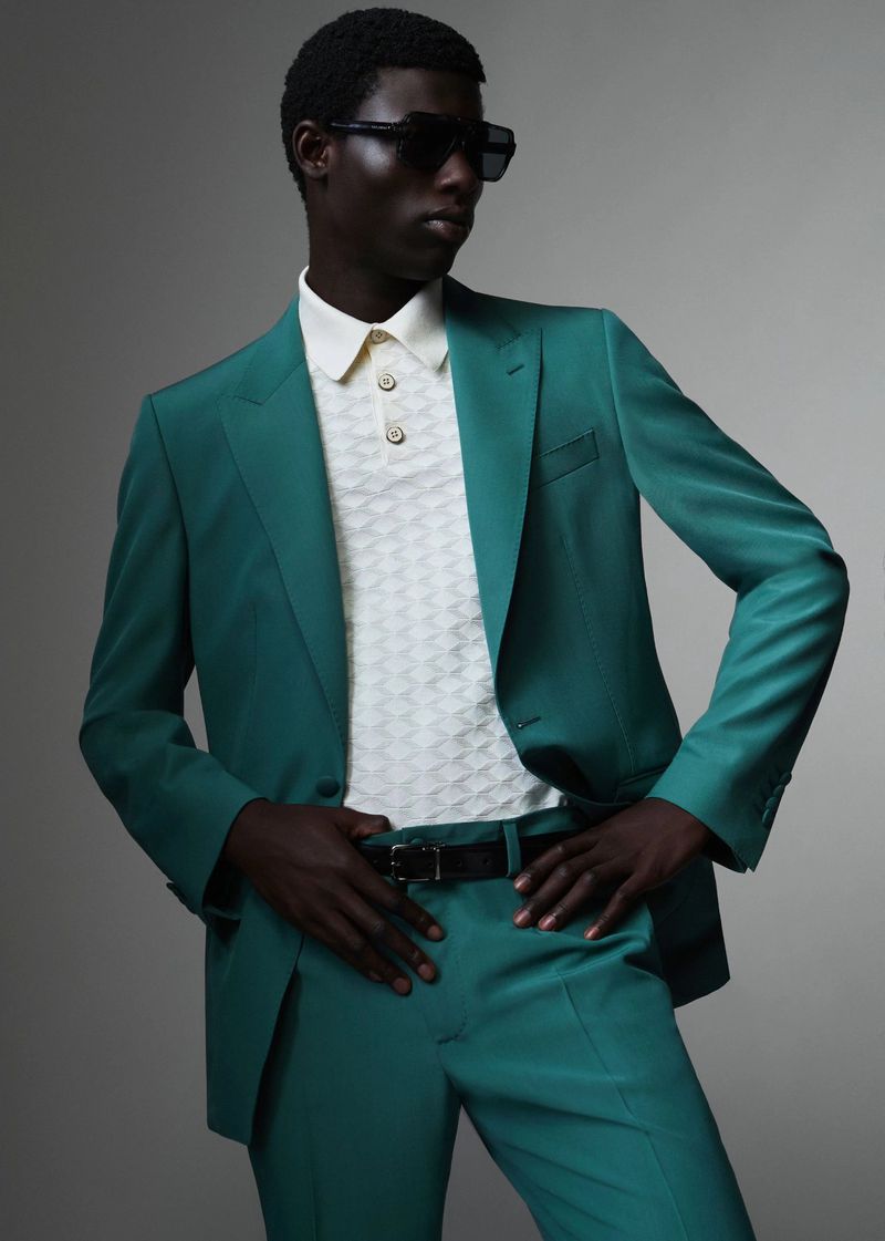 Model in emerald green suit and white polo shirt wearing dark sunglasses against a seamless grey studio background