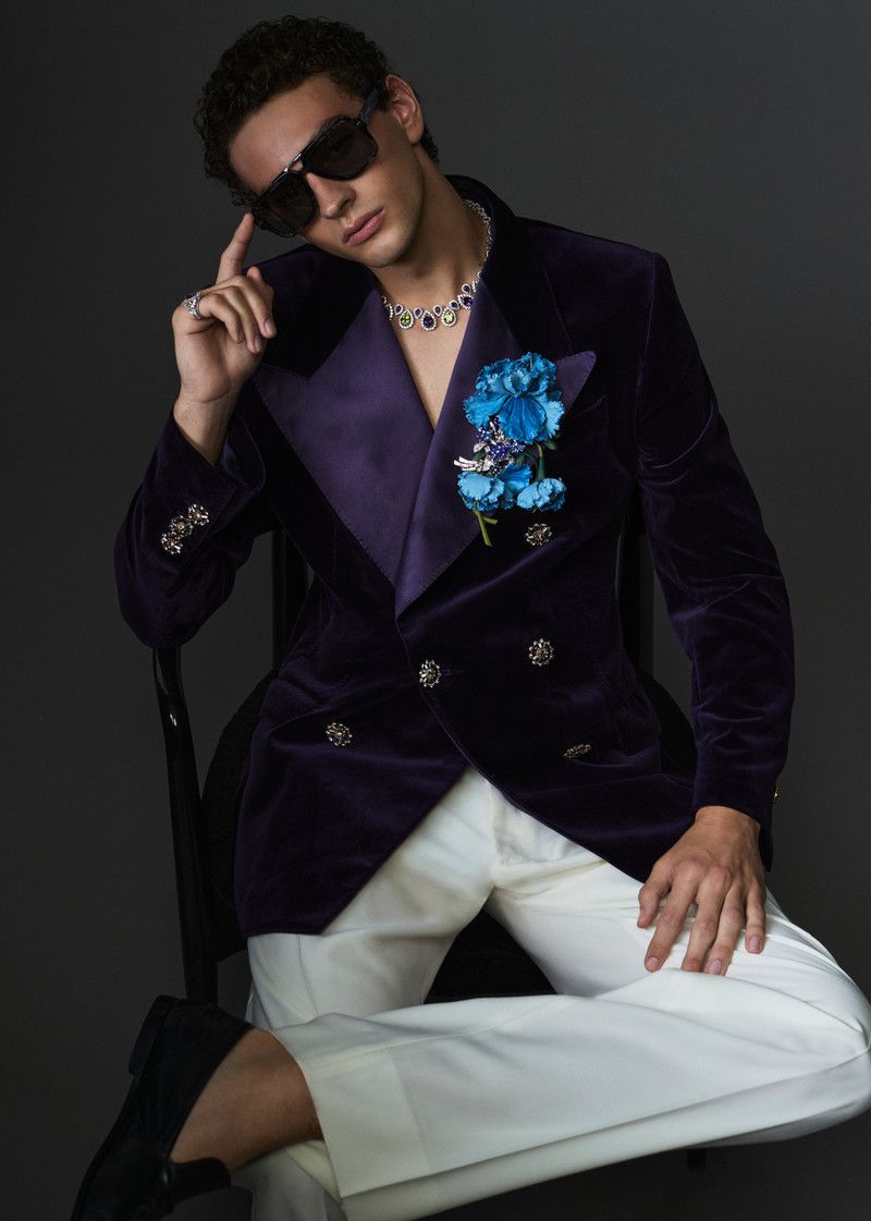 A man wearing a deep purple velvet blazer, white trousers, sunglasses, and a floral lapel pin seated against a dark grey background.