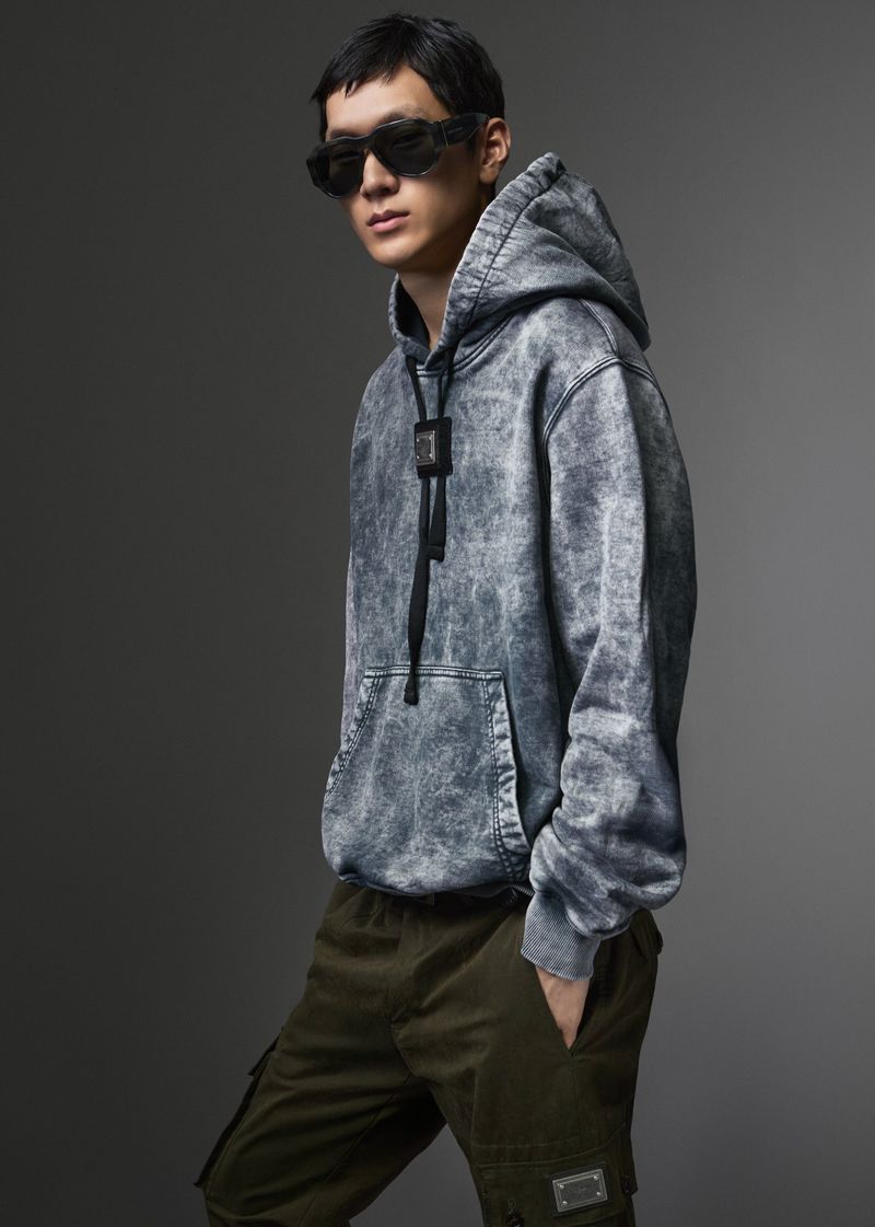Model wearing dark sunglasses, grey acid-wash hoodie, and dark cargo trousers against a neutral studio backdrop