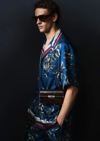 Model wearing dark sunglasses and a blue patterned silk shirt and shorts carrying a brown leather crossbody bag against a dark background.
