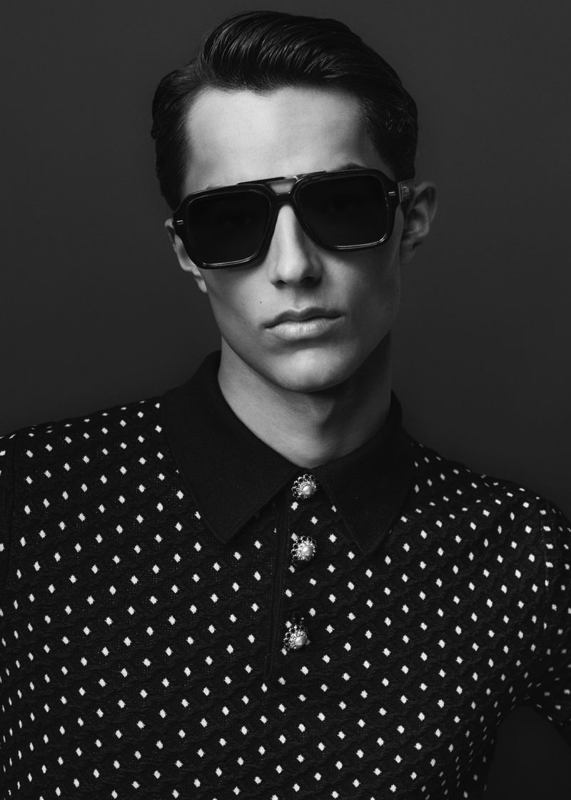 Close-up black and white portrait of a man wearing dark sunglasses and a patterned polo shirt against a dark background.