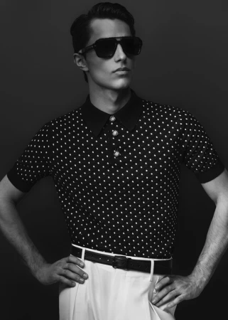 Man wearing polka dot polo shirt, white trousers, and dark sunglasses against a dark studio backdrop