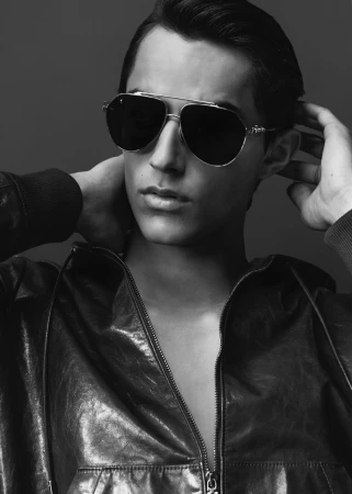 Man wearing dark sunglasses and a zipped leather jacket posing against a plain grey background