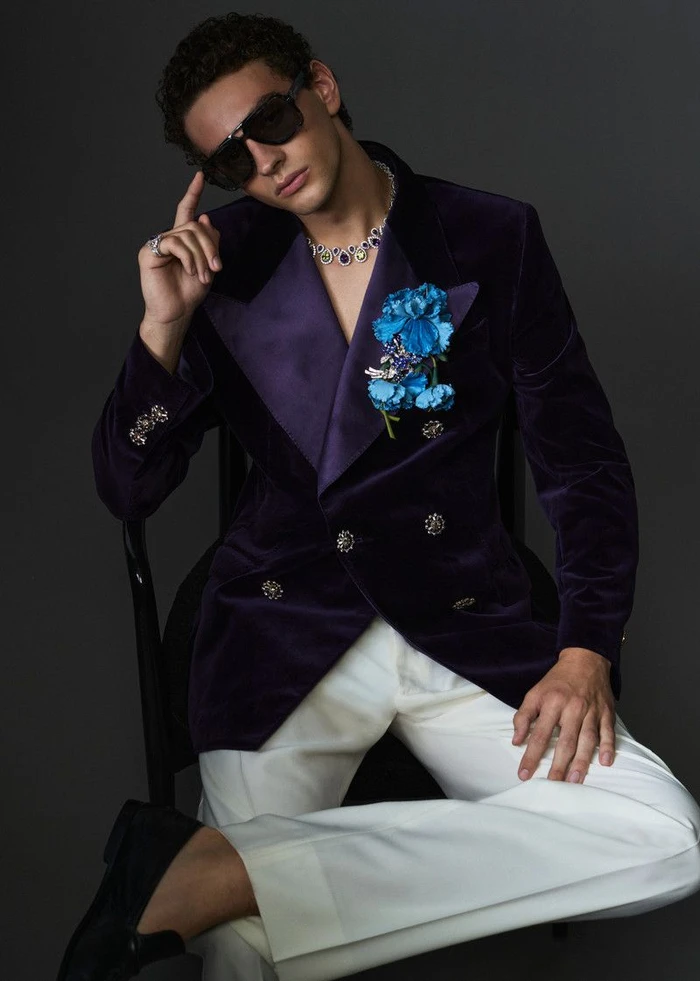 A man wearing a deep purple velvet blazer, white trousers, sunglasses, and a floral lapel pin seated against a dark grey background.
