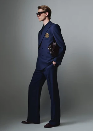 Full-length portrait of a male model wearing a navy blue double-breasted suit and sunglasses against a grey studio backdrop.