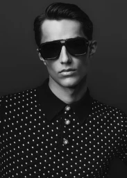 Close-up black and white portrait of a man wearing dark sunglasses and a patterned polo shirt against a dark background.