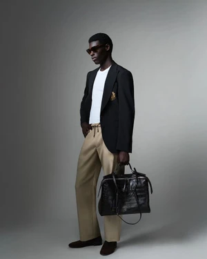 Man wearing a black blazer over a white t-shirt and tan trousers holding a large black leather bag against a grey background.