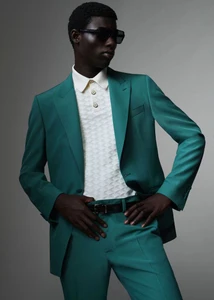 Model in emerald green suit and white polo shirt wearing dark sunglasses against a seamless grey studio background
