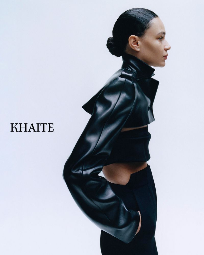 Profile view of a woman in black leather jacket and pants against a white backdrop, showcasing modern fashion and minimal styling.