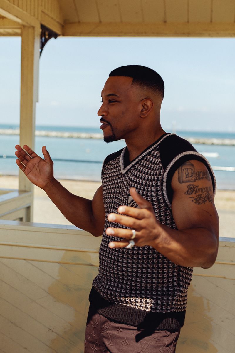 Medium shot of an adult man with short hair and a goatee, wearing a patterned sweater vest and patterned pants, standing outside by the water.