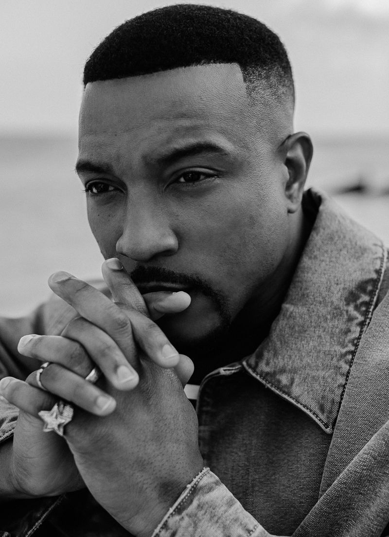 Black and white portrait of an adult male with hands clasped near his face, wearing a denim jacket with a body of water background.