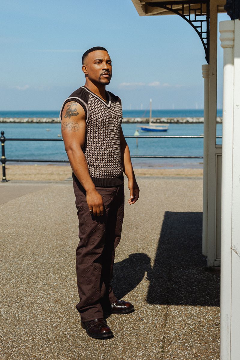 Portrait of a muscular adult male wearing a patterned sleeveless sweater and brown pants, standing by a white structure near the ocean.