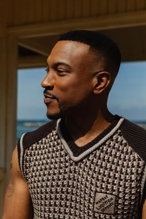 Close-up of an adult male with short dark hair, wearing a patterned vest, looking off to the side in an outdoor setting with natural light.