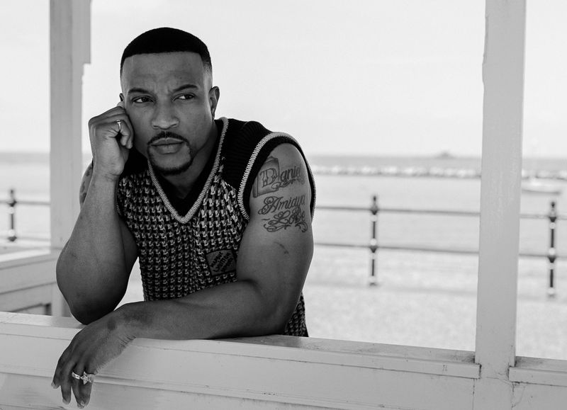 Portrait of a dark-skinned adult man in a patterned vest, leaning on a white structure with the ocean in the background.