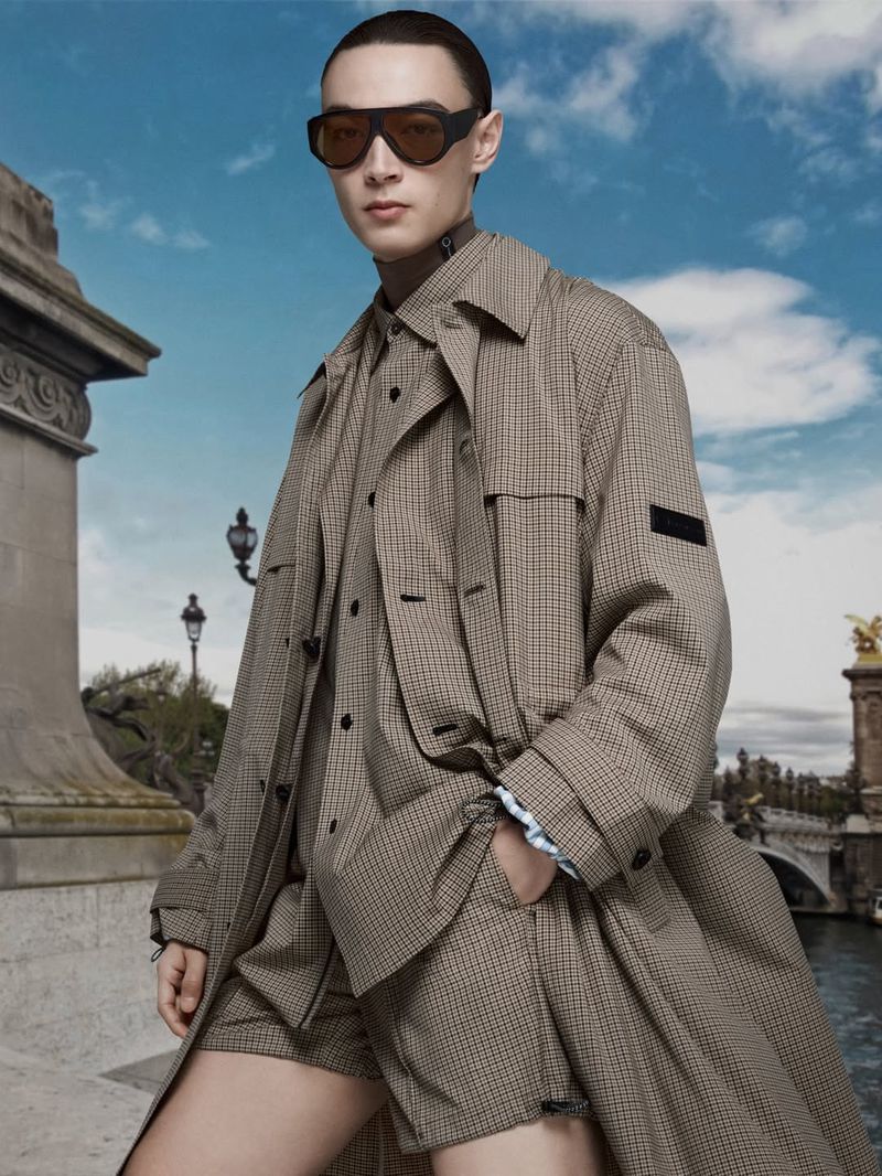 Model wearing dark sunglasses and a patterned beige coat outdoors against a bridge and cloudy blue sky background