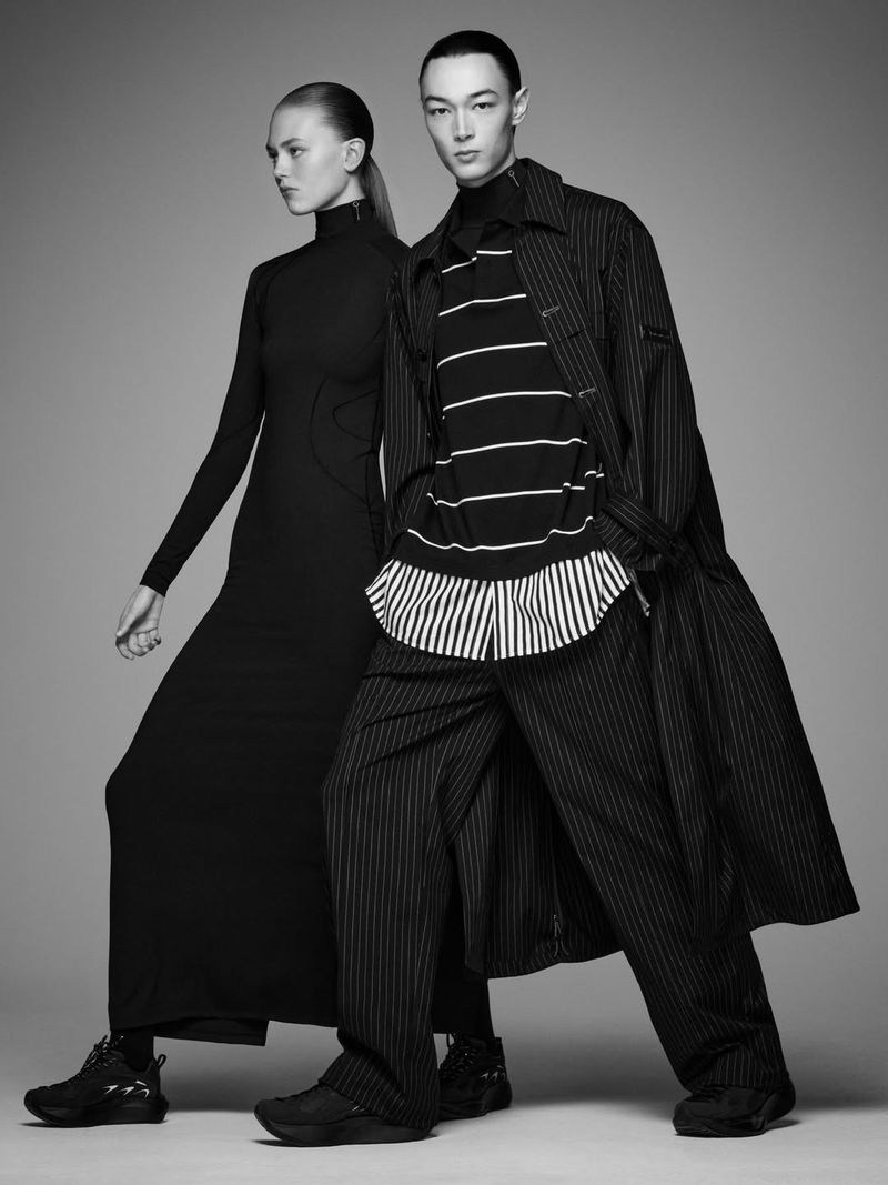 Two fashion models in black and white studio. The woman wears a long dress, while the man has a striped shirt and overcoat.
