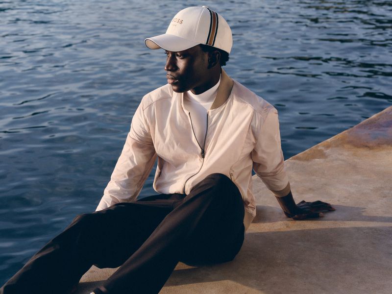 Man sitting on a concrete ledge by the water wearing a cream bomber jacket, white turtleneck, and Boss cap.