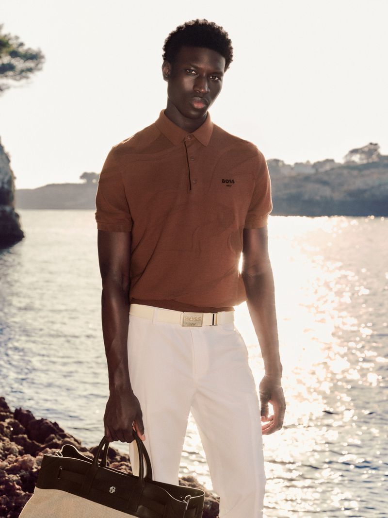 A stylish Black man in a brown polo shirt and white pants poses outdoors on a rocky shore holding a leather tote bag.