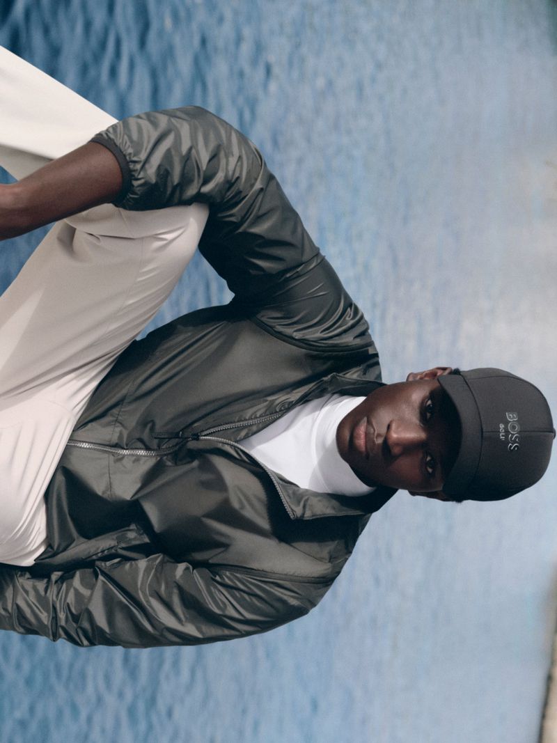 Black man wearing dark windbreaker jacket, white turtleneck, and black cap outdoors near water