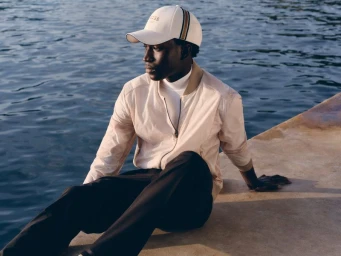 Man sitting on a concrete ledge by the water wearing a cream bomber jacket, white turtleneck, and Boss cap.