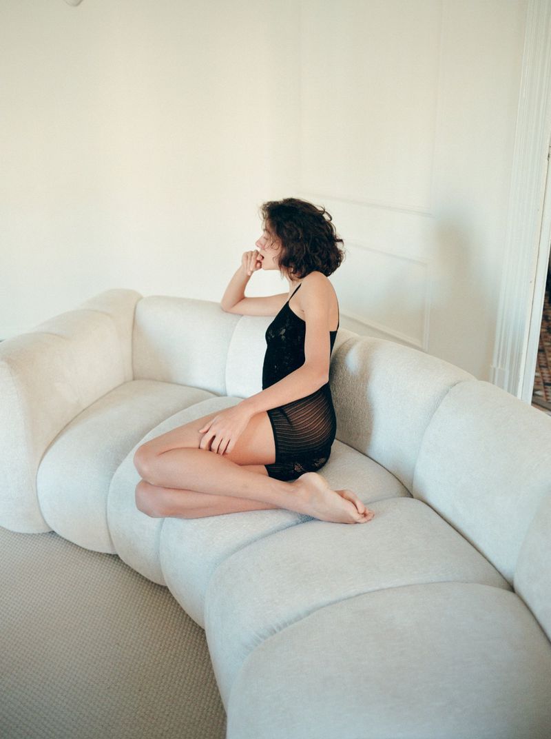 Woman with short dark curly hair wearing a sheer black slip dress seated on a large white sectional sofa indoors.
