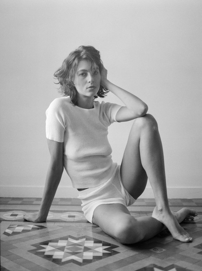 Woman in white top and shorts sitting on patterned tile floor against a plain wall