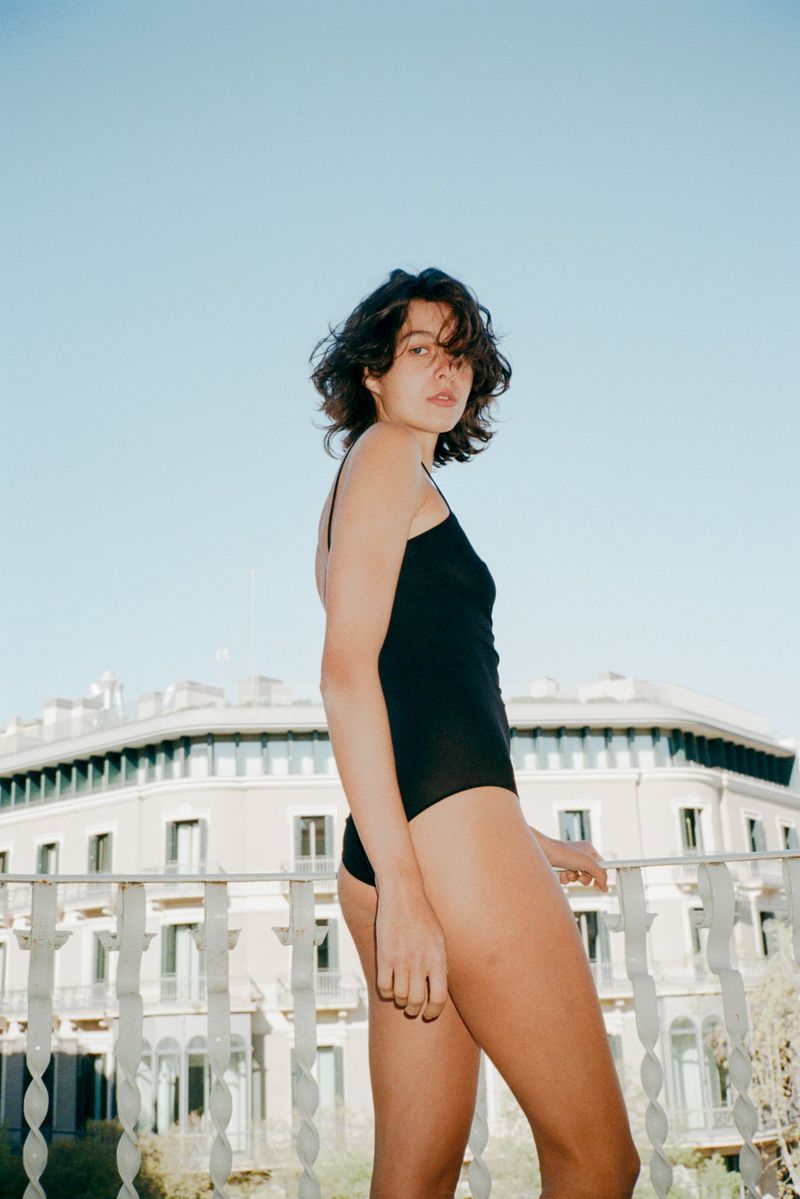 Woman in a black bodysuit leaning on a white balustrade against an exterior building under a clear blue sky