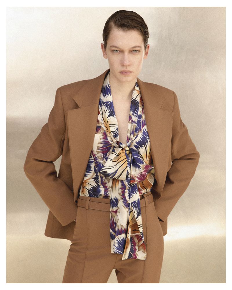 Model in brown tailored suit jacket and matching trousers with a patterned silk bow blouse against a neutral textured background