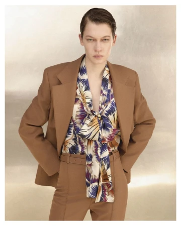 Model in brown tailored suit jacket and matching trousers with a patterned silk bow blouse against a neutral textured background