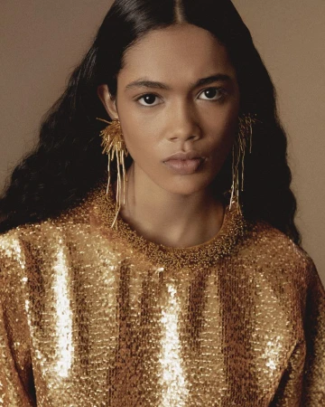 Close-up portrait of a model wearing a shimmering gold sequin top and intricate fringed gold earrings against a neutral background.