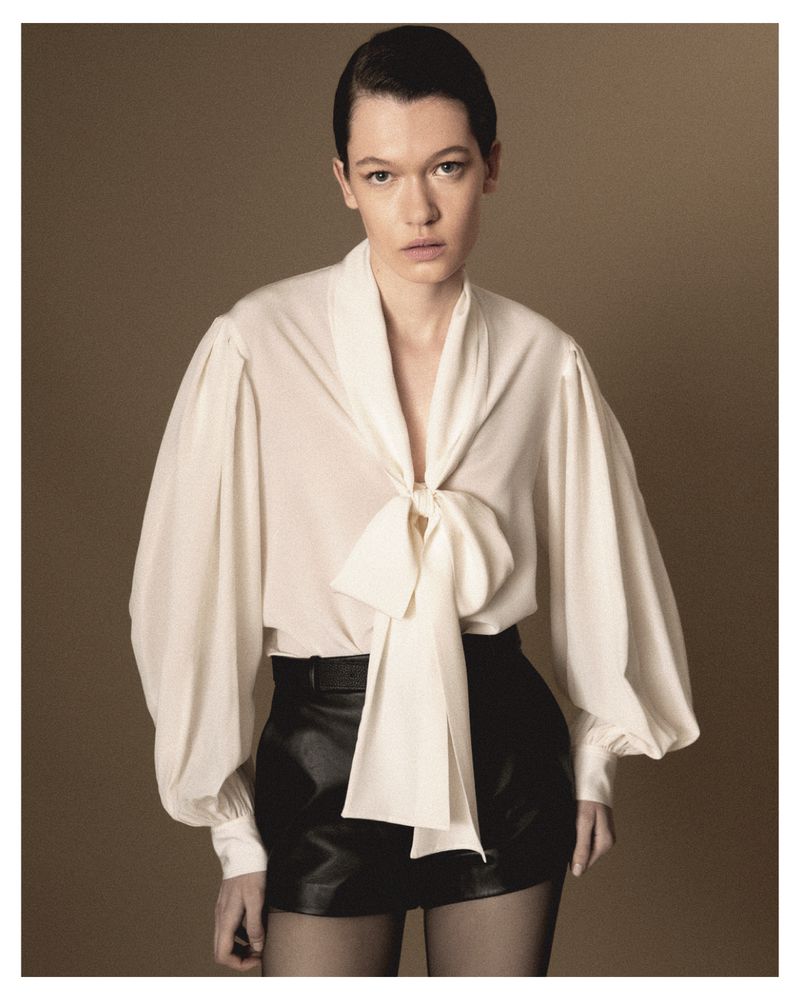 Model wearing a white tie-neck blouse with balloon sleeves and black shorts against a solid brown studio background.
