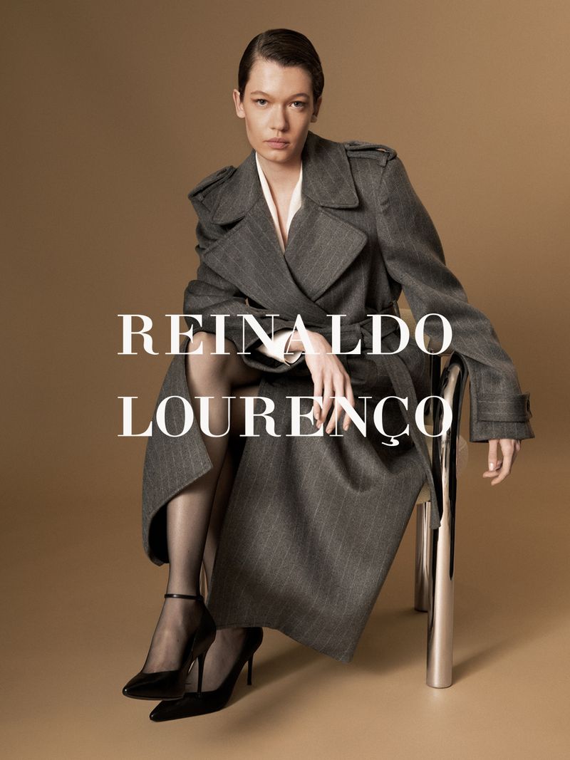 Model seated on a chrome chair wearing a charcoal grey trench coat against a solid warm background.