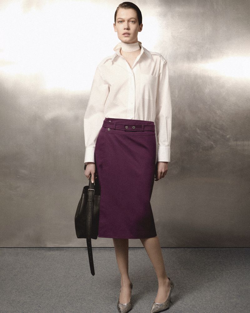 Model wearing a white shirt and purple skirt holding a handbag against a textured metallic background