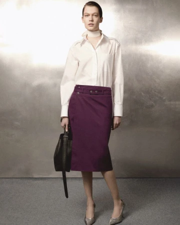 Model wearing a white shirt and purple skirt holding a handbag against a textured metallic background