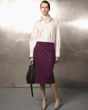 Model wearing a white shirt and purple skirt holding a handbag against a textured metallic background