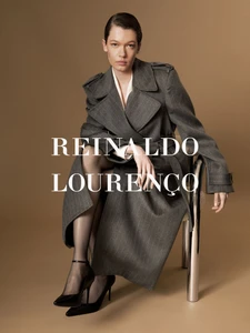 Model seated on a chrome chair wearing a charcoal grey trench coat against a solid warm background.
