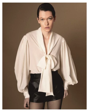 Model wearing a white tie-neck blouse with balloon sleeves and black shorts against a solid brown studio background.