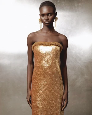 Model wearing a strapless metallic gold sequin dress and fringed earrings against a neutral textured background