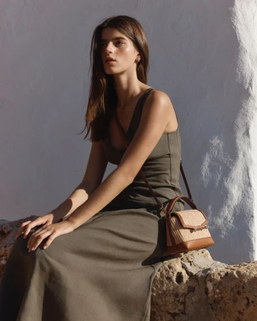 Model wearing olive green dress holding leather straw handbag sitting outdoors against white wall