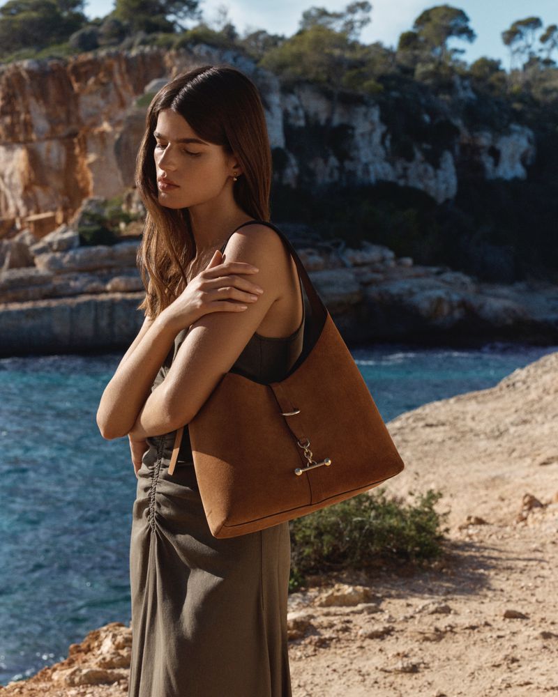 Model wearing a dark green dress holding a brown suede handbag by the rocky coast for Strathberry High-Summer Collection 2026