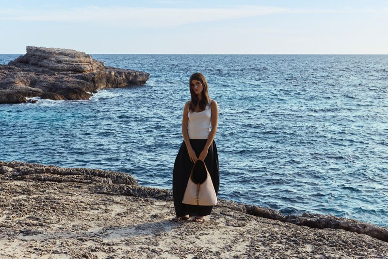 Model standing on a rocky coast holding a light leather handbag with the blue ocean behind her
