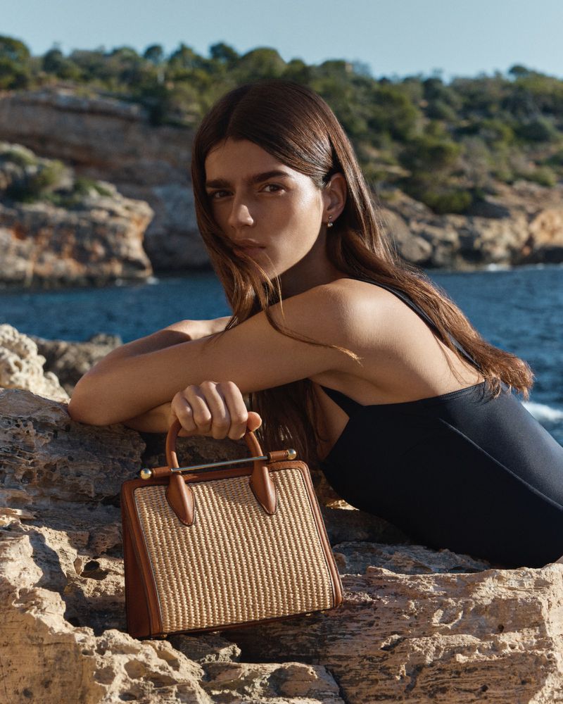 Woman leaning on rocky coastal terrain holding a straw handbag with leather trim against an ocean backdrop