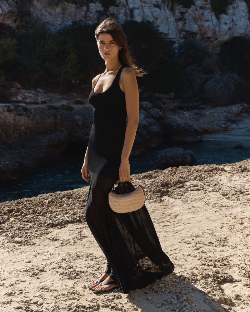 Model wearing a long black dress holding a woven handbag on a sandy beach