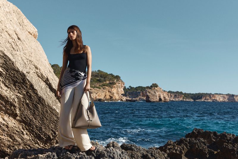 Model standing on rocky coastal shore holding a large leather shoulder bag against ocean and cliffs