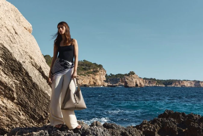 Model standing on rocky coastal shore holding a large leather shoulder bag against ocean and cliffs