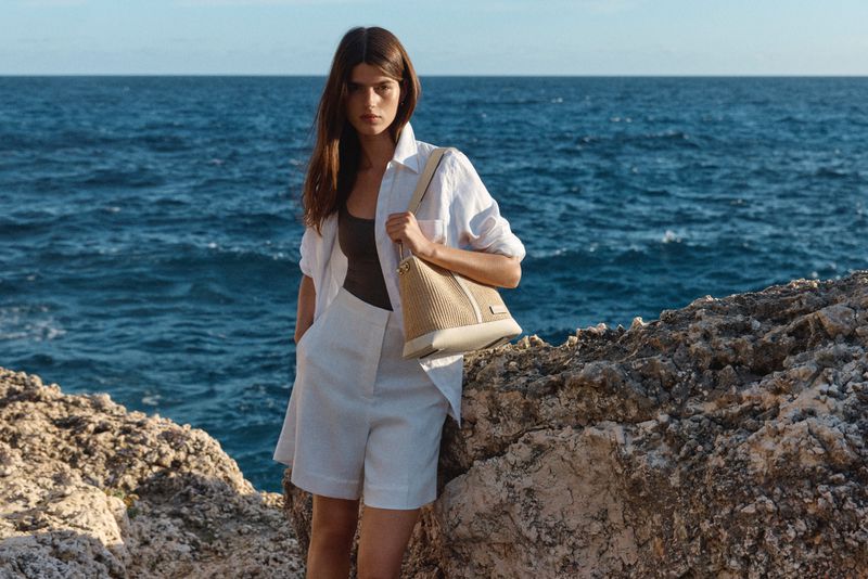 Model wearing a white shirt and shorts holding a straw handbag against rocky coastline and ocean background