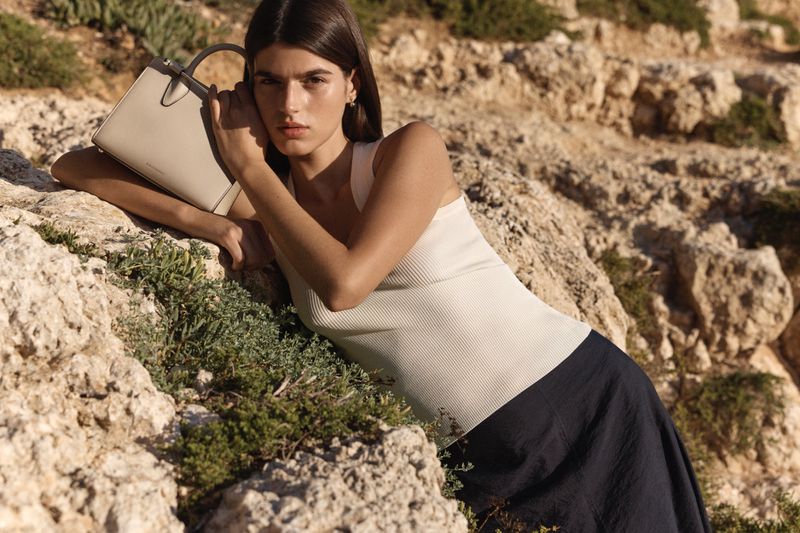 Model leaning on rocky coastal terrain wearing a white tank top and holding a structured handbag