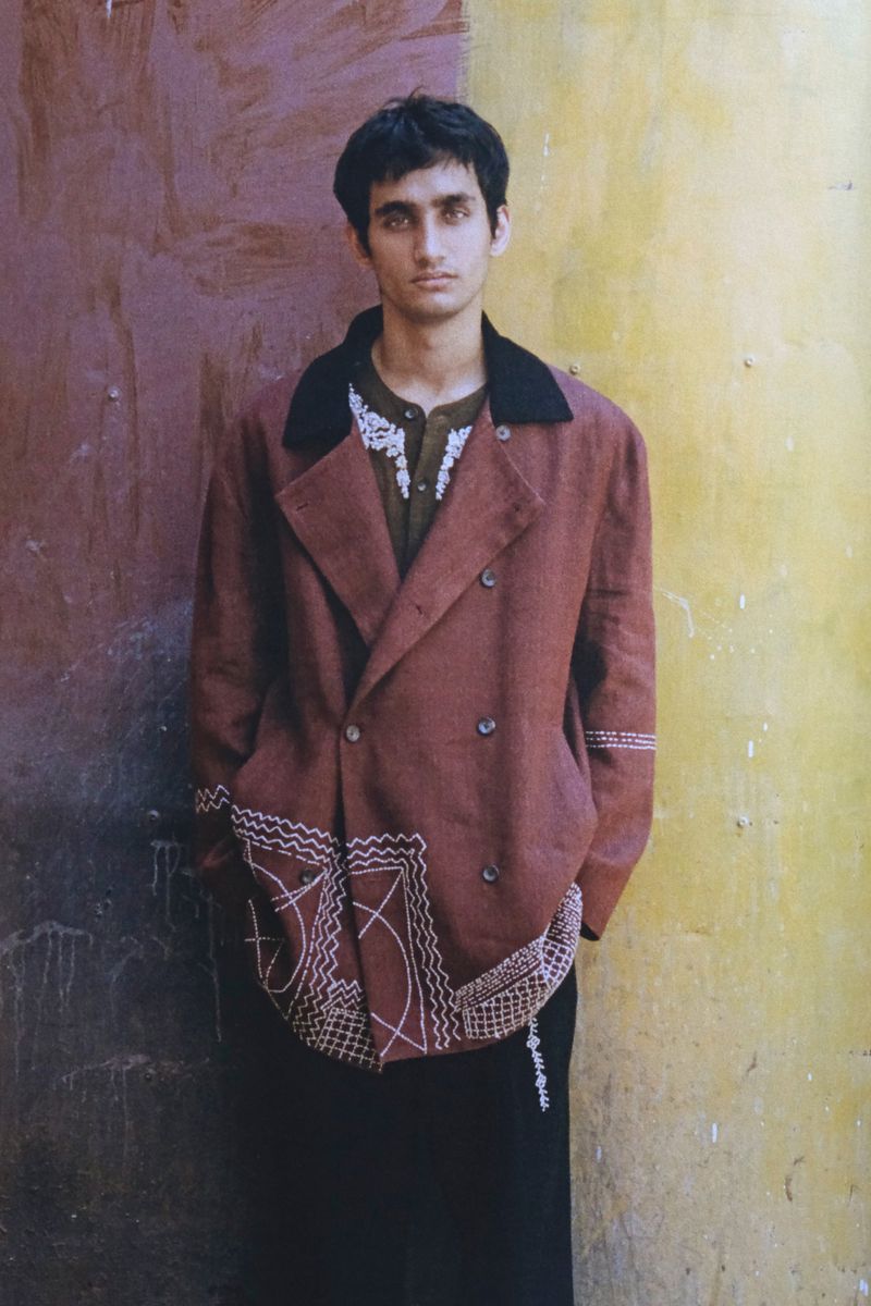 Man wearing a brown double-breasted jacket with white embroidery standing against a multicolored wall