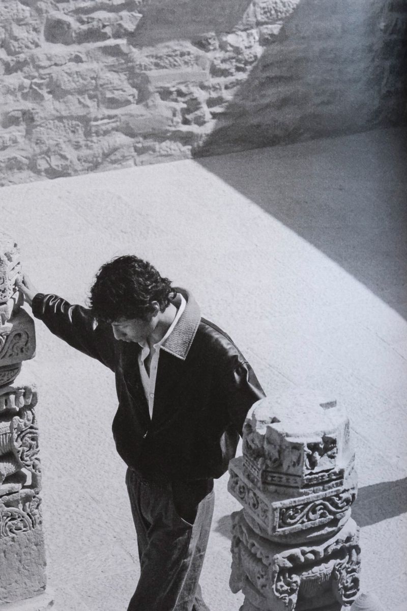 Monochrome image of a man in a jacket leaning against an ornate stone pillar, with a textured stone wall and floor in the background.